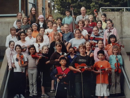 A group of young string players stand on a stair case smiling for the camera.