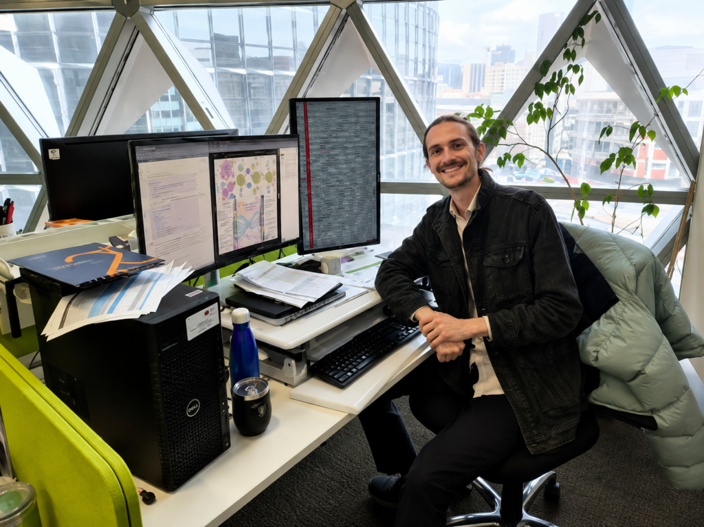 A man sits at his desk, turned smiling to the camera, in an office environment.